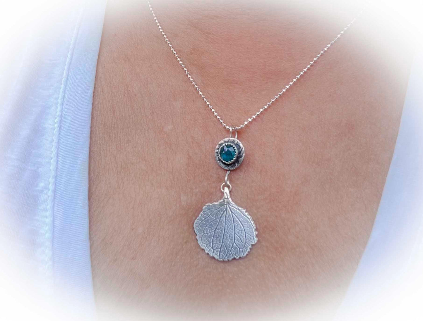 Silver necklace with a leaf pendant and blue gemstone on a person's neck.
